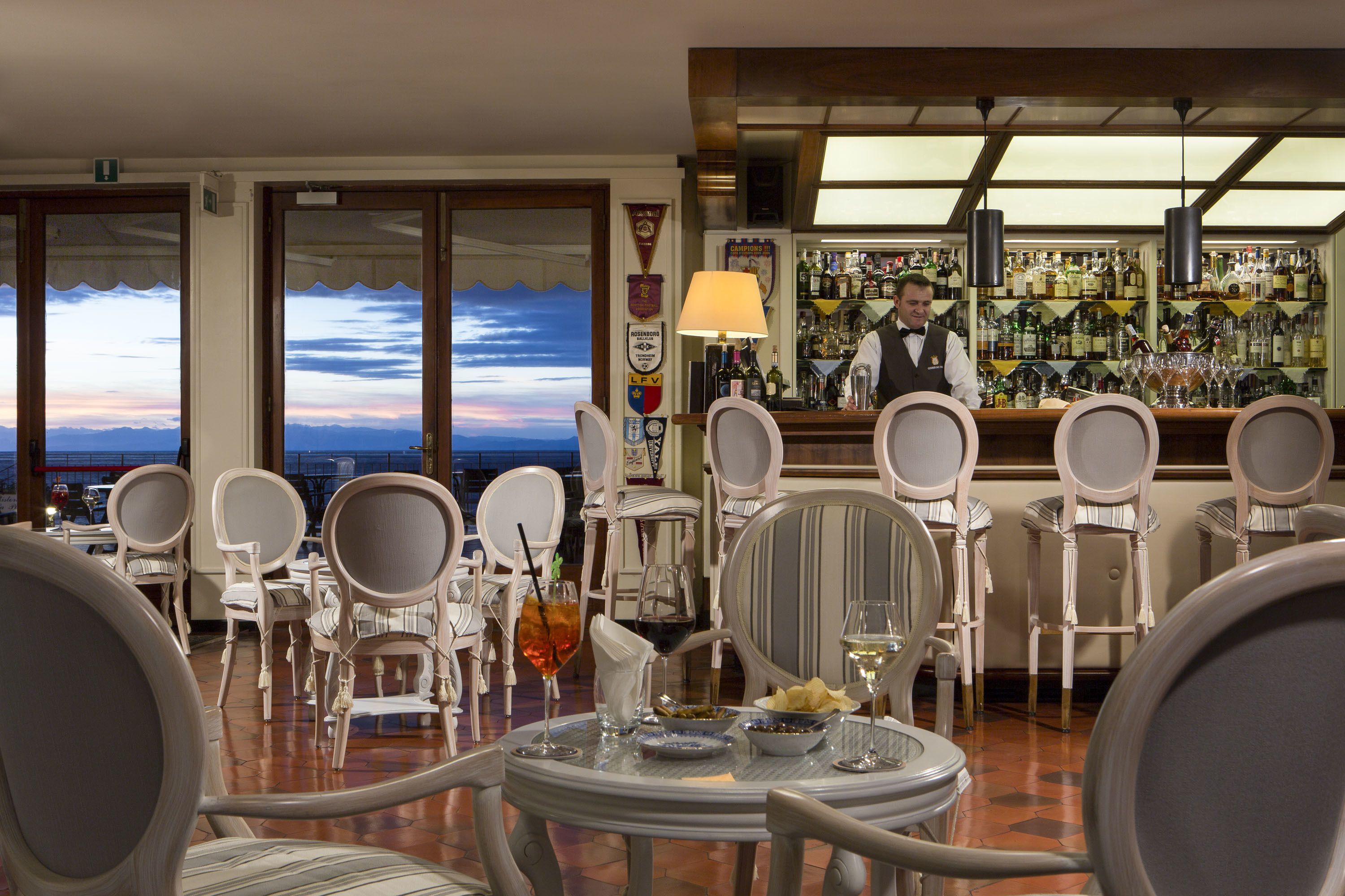 Un área de bar con un bartender, vista del cielo a través de amplios ventanales y varias mesas con sillas.