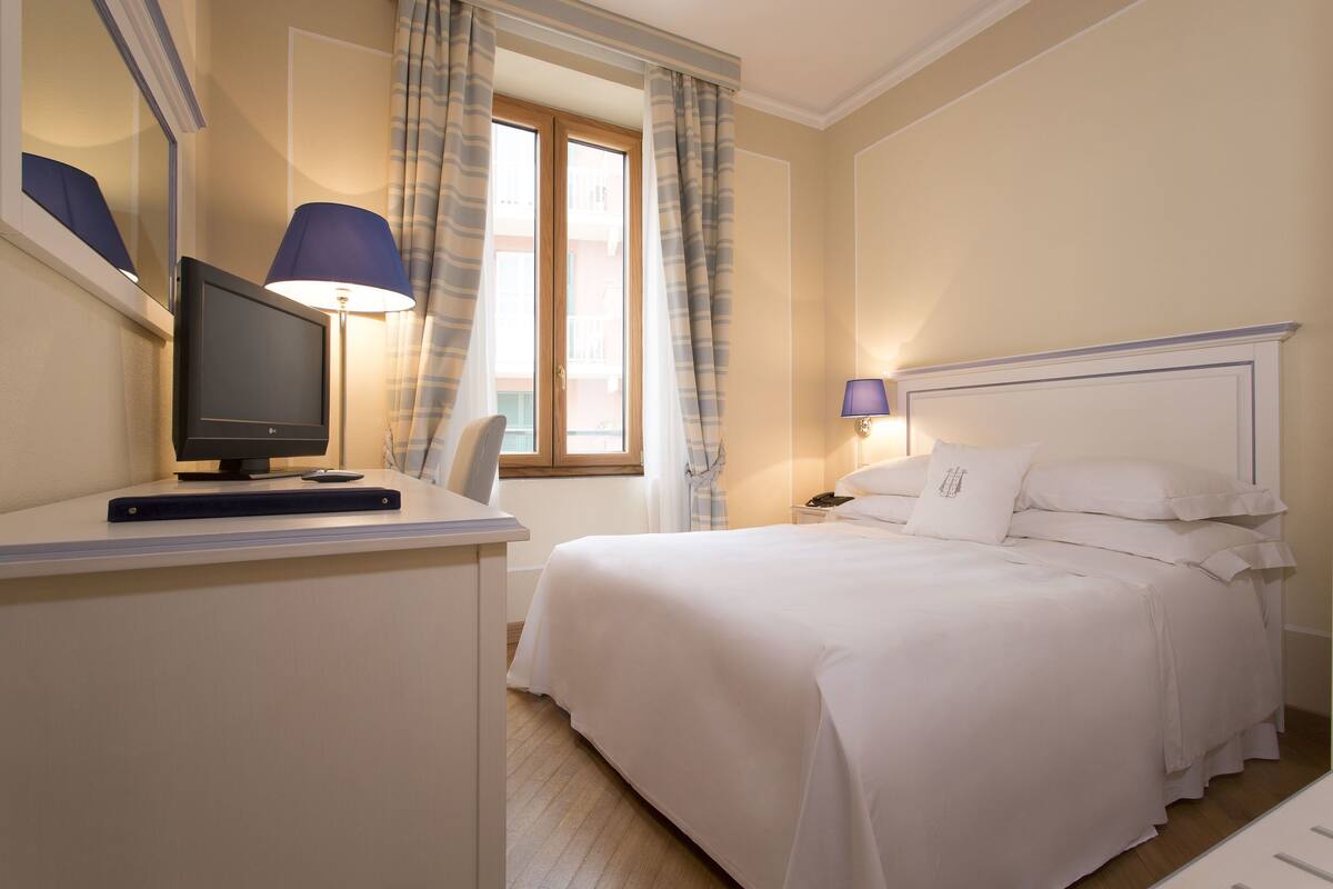 Une chambre d’hôtel comprenant un lit, un bureau avec un ordinateur, une chaise, une fenêtre avec des rideaux et une table de chevet avec une lampe.