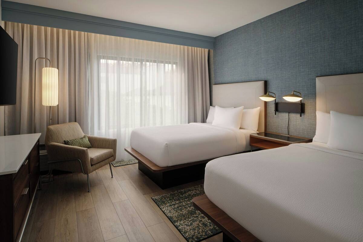 Habitación de hotel con dos camas, un escritorio, una silla, un televisor y un ventanal con cortinas.