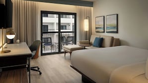 A hotel room with a bed, desk, chair, and a balcony with a view of the building. - Courtyard by Marriott Orlando Airport (Orlando)