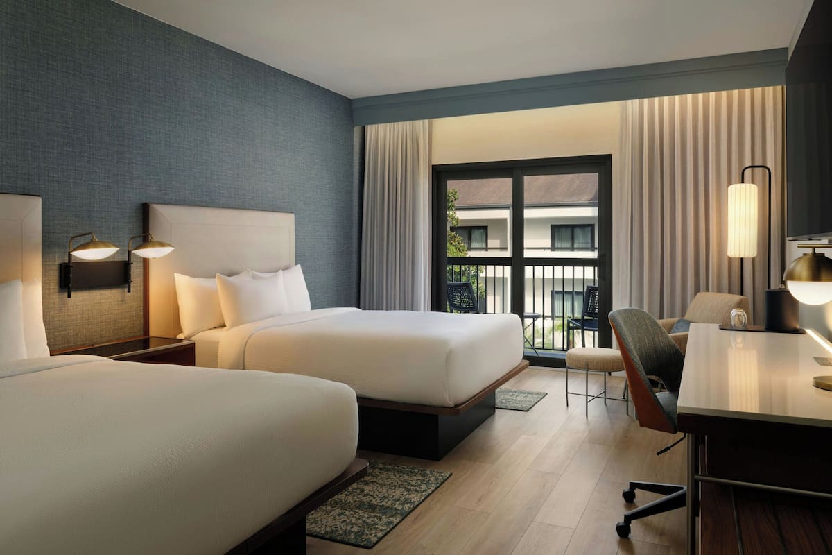 Een hotelkamer met twee bedden, een bureau, een stoel en een balkon met gordijnen.