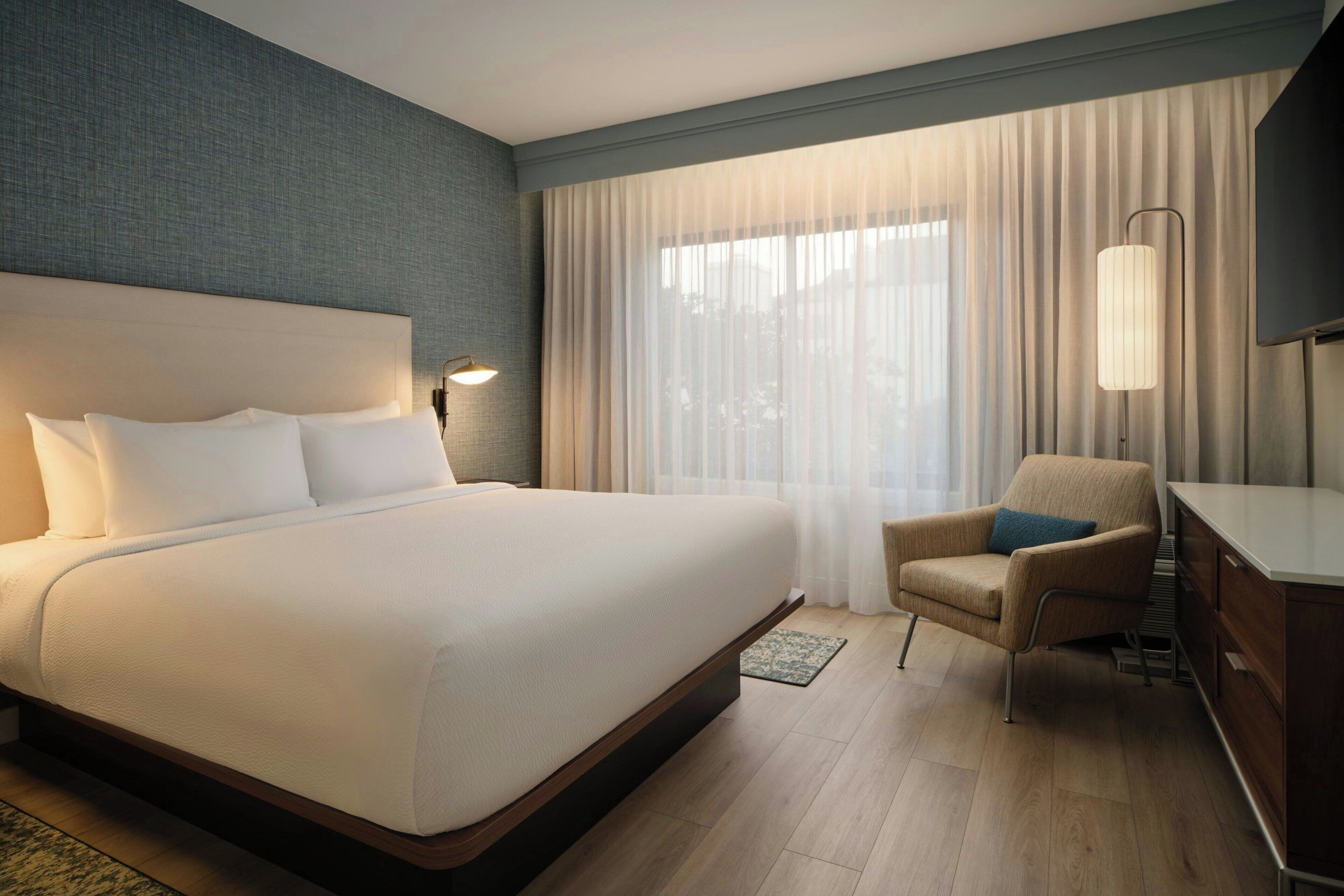 A hotel room with a large bed, a chair, a desk, a lamp, and a window with curtains.