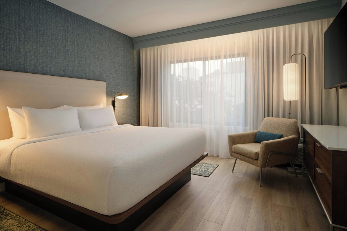 A hotel room with a large bed, a chair, a desk, a lamp, and a window with curtains.