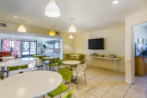 A modern dining area with round tables and green chairs, a flat-screen TV, and a kitchen area visible through an open door.