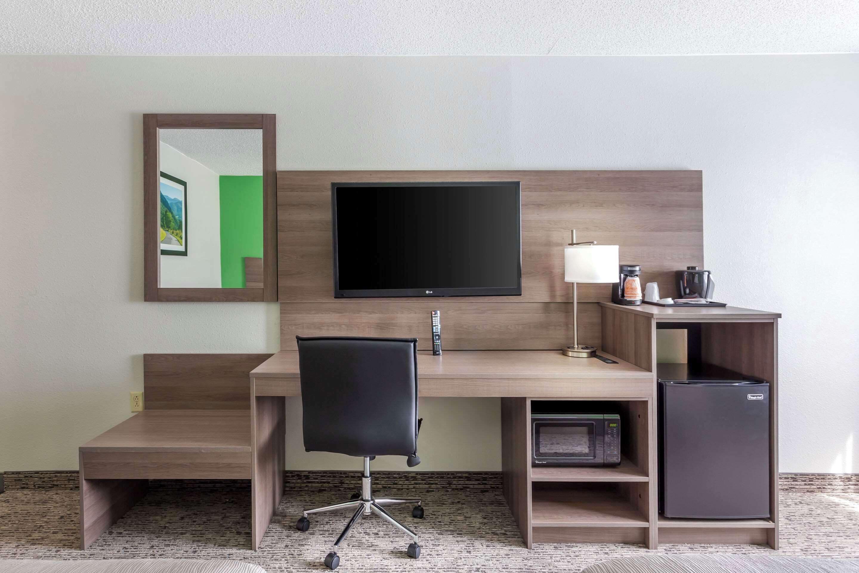 A hotel room with a wooden desk, a black office chair, a flat-screen TV, a lamp, a coffee maker, and a mini-fridge.