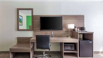 A hotel room with a wooden desk, a black office chair, a flat-screen TV, a lamp, a coffee maker, and a mini-fridge.