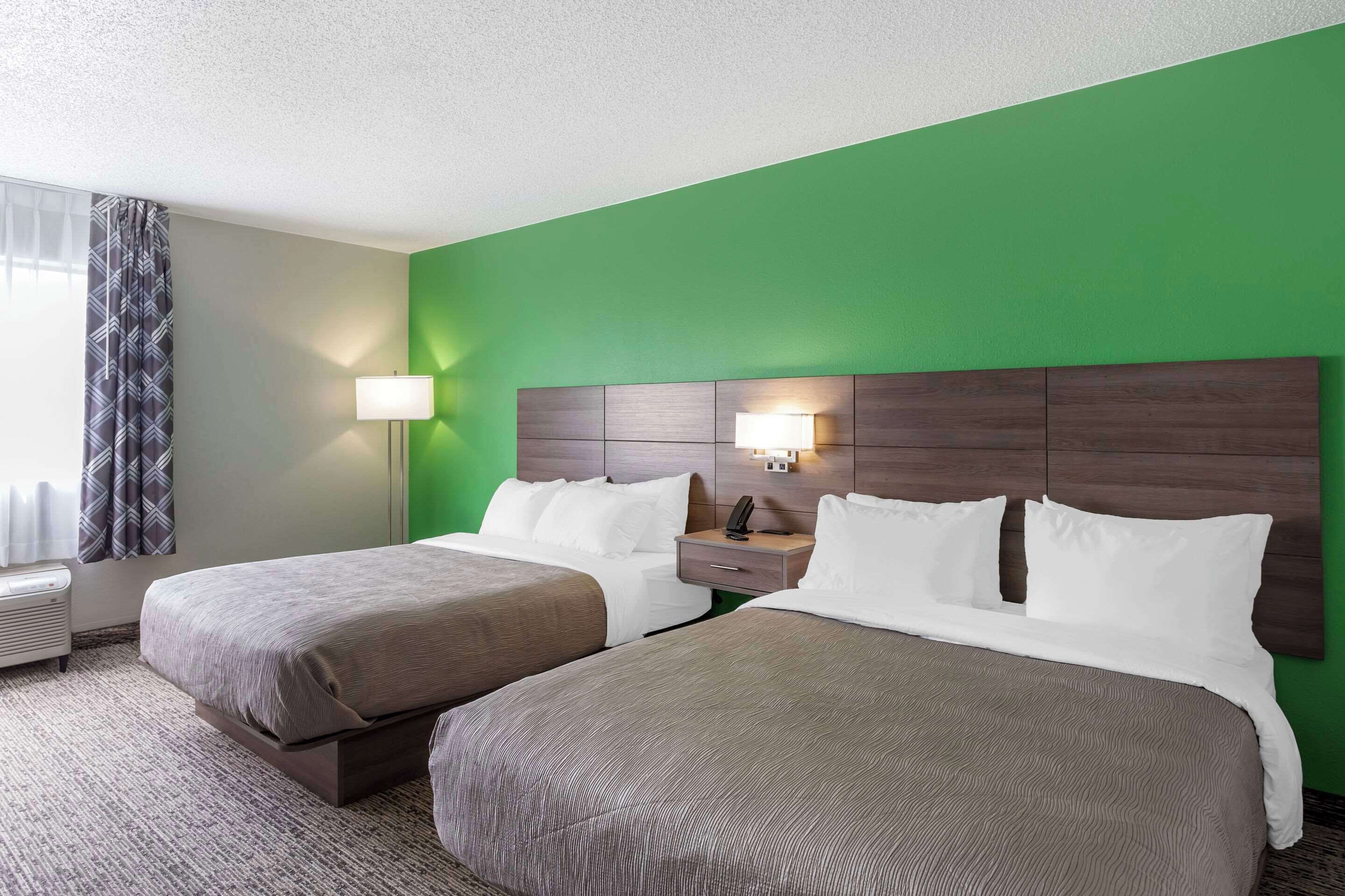 A hotel room with two beds, a green accent wall, a nightstand with a lamp, and a window with curtains.