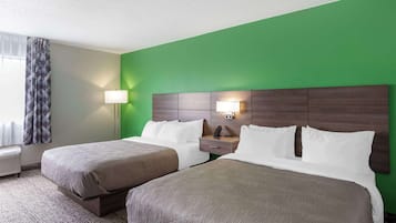 A hotel room with two beds, a green accent wall, a nightstand with a lamp, and a window with curtains.