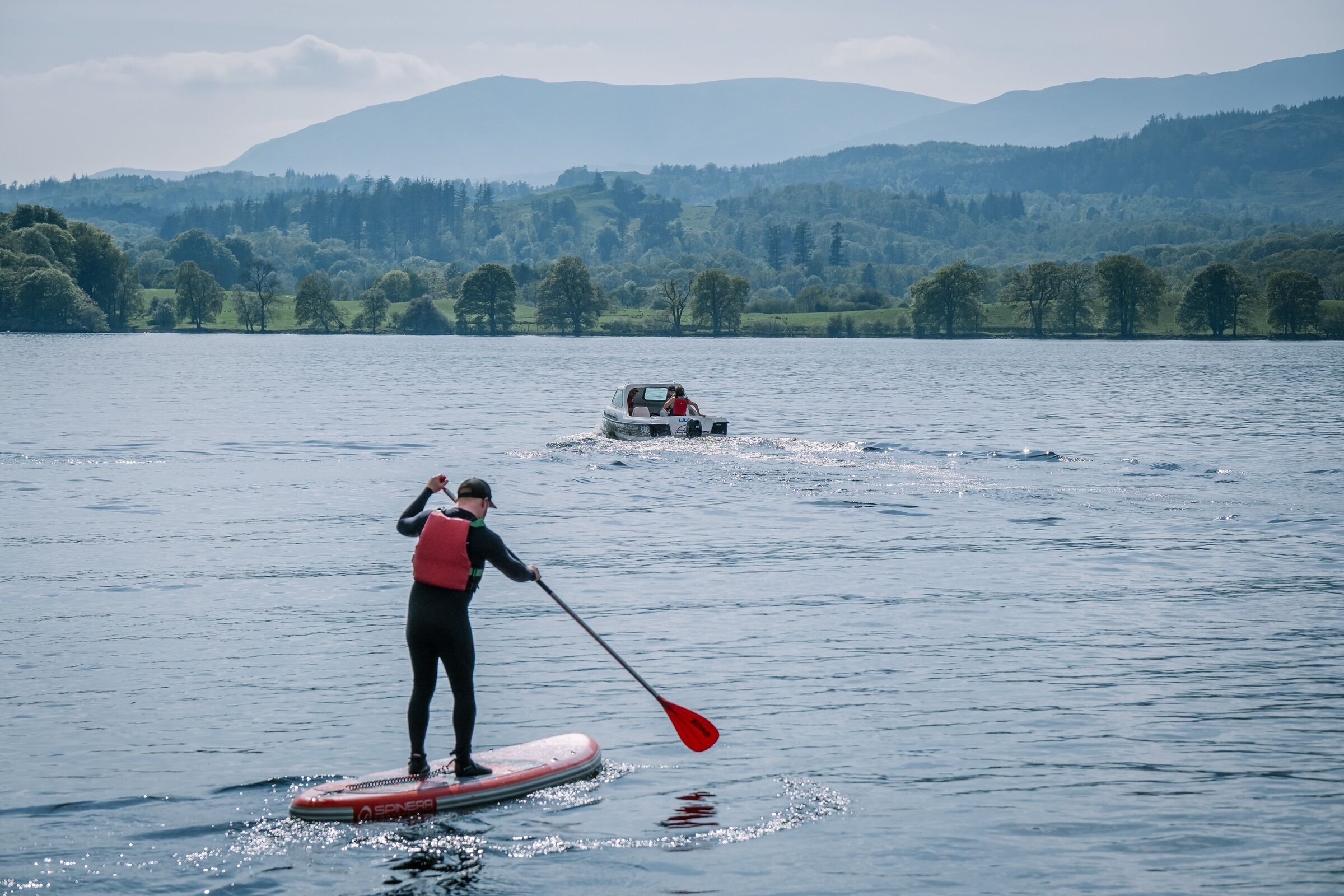 A person paddleboarding on a calm lake with a boat in the background and hills in the distance.