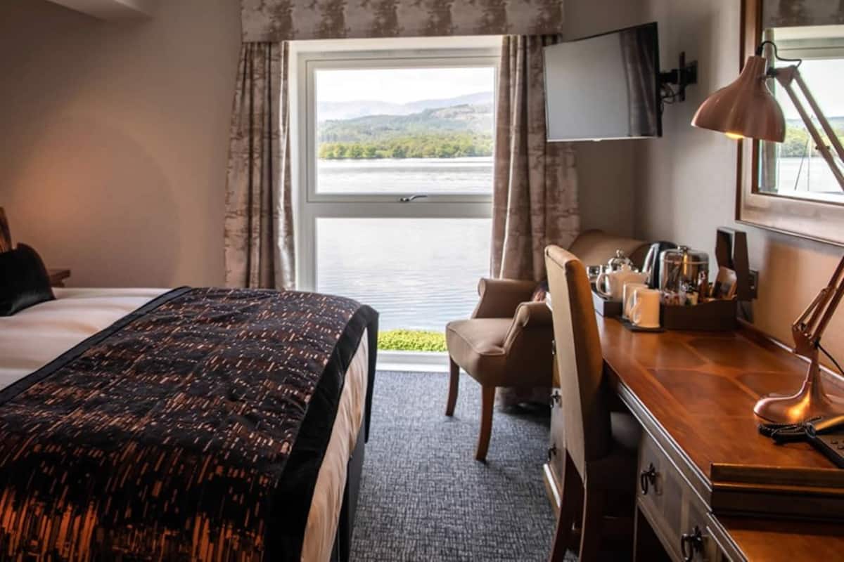 A hotel room with a bed, a desk, a chair, a desk lamp, and a view of a lake through a window.