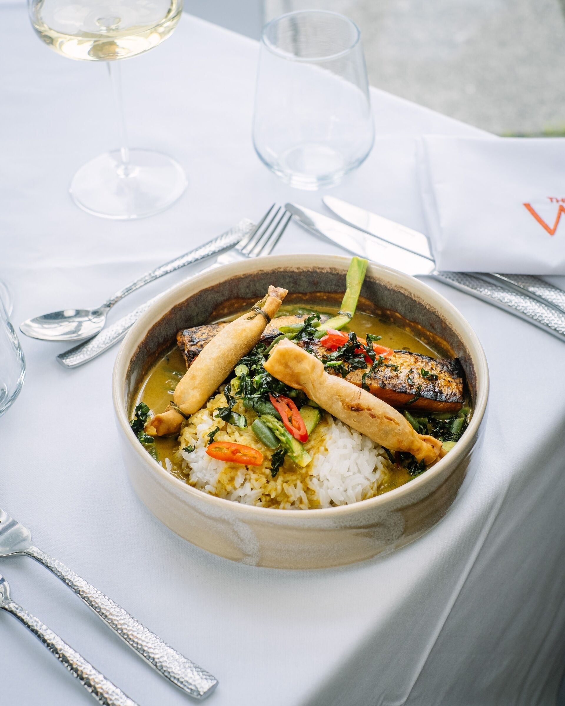 A bowl of food with rice, vegetables, and a piece of meat, accompanied by cutlery and a glass of white wine on a table.