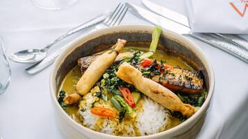 A bowl of food with rice, vegetables, and a piece of meat, accompanied by cutlery and a glass of white wine on a table.