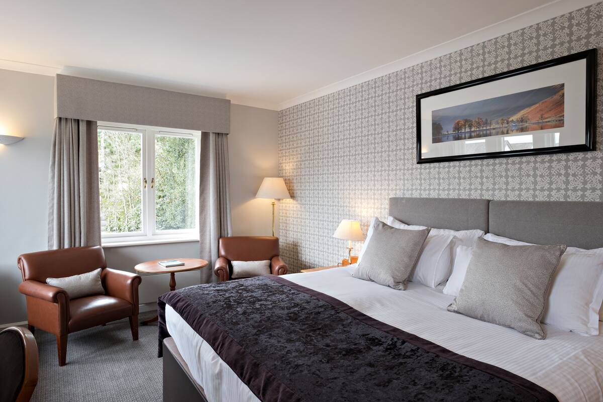 A hotel room with a large bed, two armchairs, a small table, a lamp, and a framed picture on the wall.