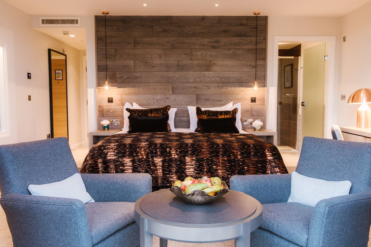 A modern bedroom with a large bed, two nightstands, a round table with a fruit bowl, and two armchairs.