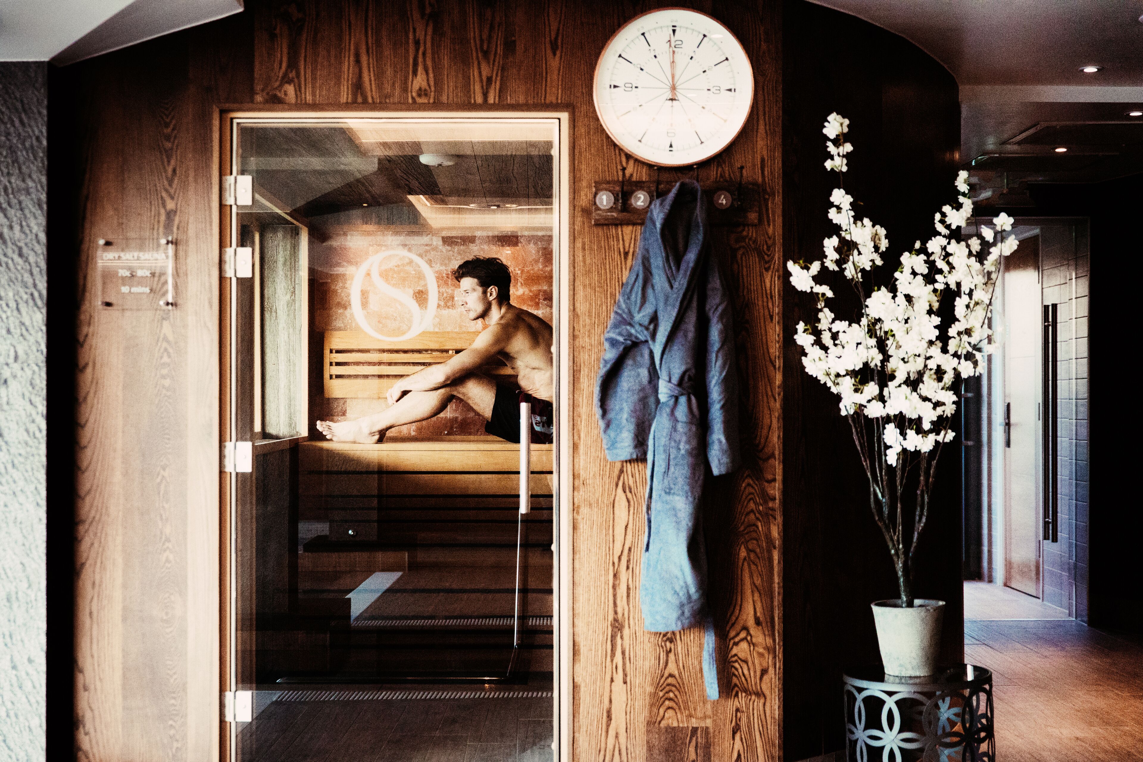Een houten interieur met een glazen deur naar een sauna, een klok en een potplant.