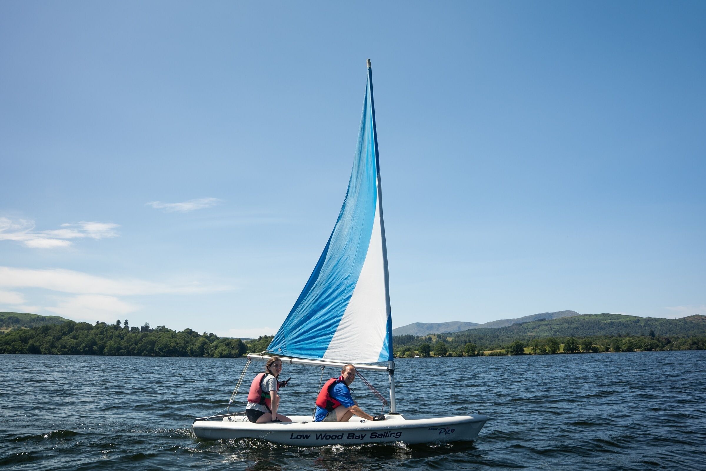 Un voilier avec deux personnes à bord, naviguant sur un lac avec des collines en arrière-plan.