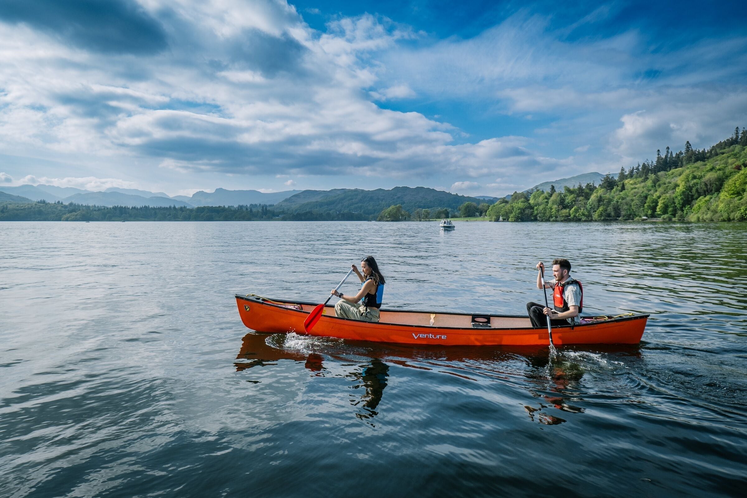 Un canoë rouge sur une étendue d’eau calme, avec deux personnes qui pagayent, entouré d ’un paysage boisé et de montagnes à l’horizon.