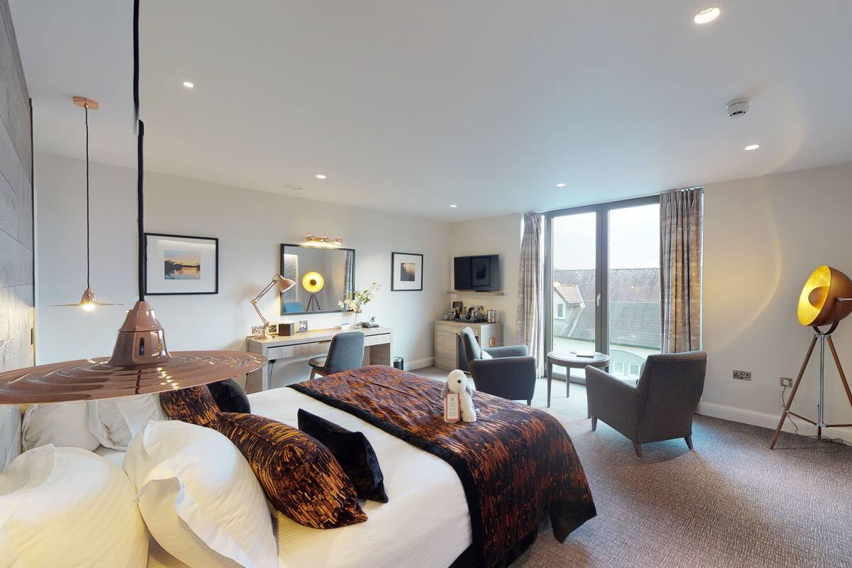 A modern hotel room with a large bed, a desk, a chair, a TV, and a large window.