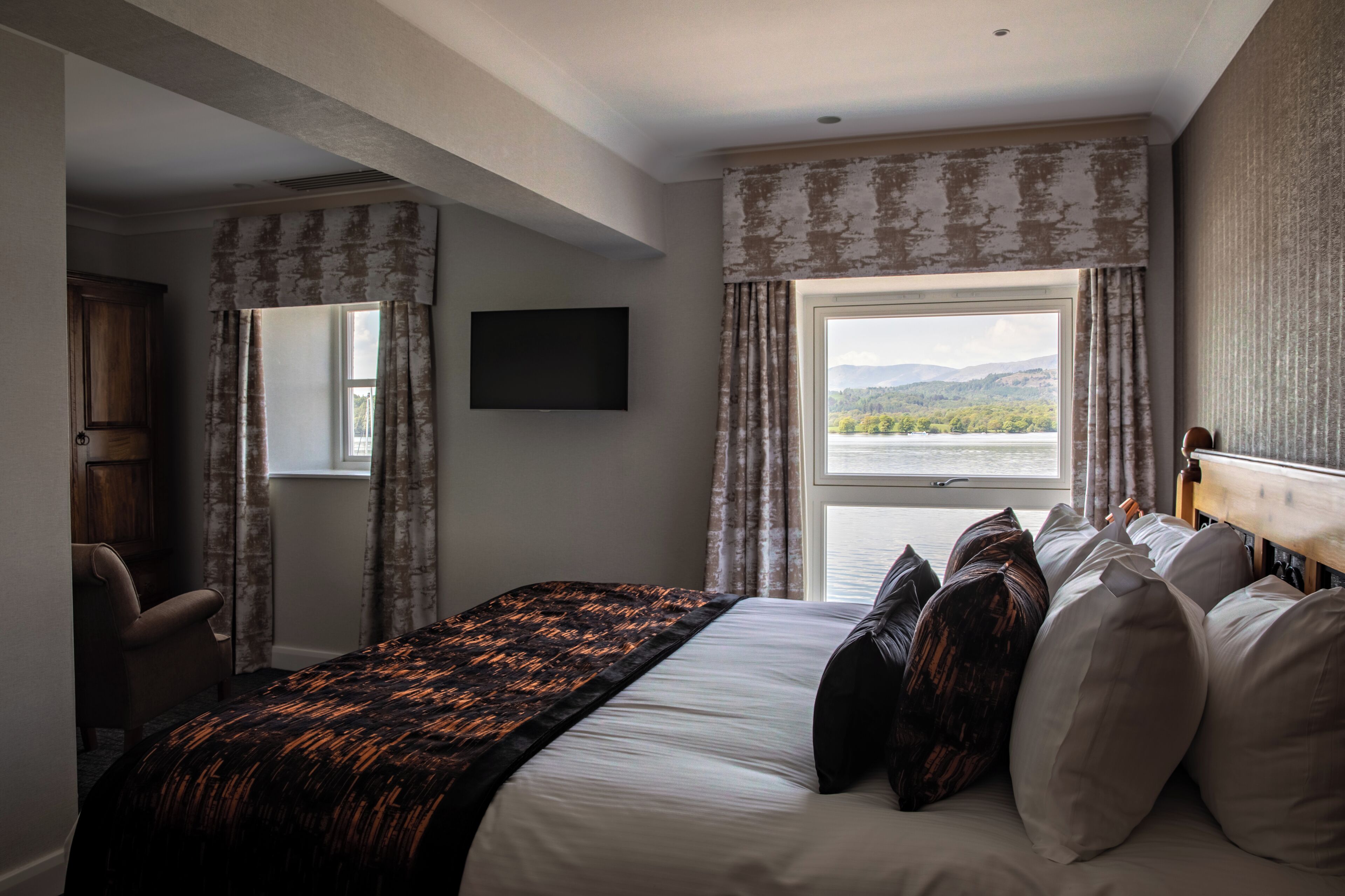 Una camera d'albergo con un letto, una televisione, una finestra con tende e vista su un lago e delle montagne.