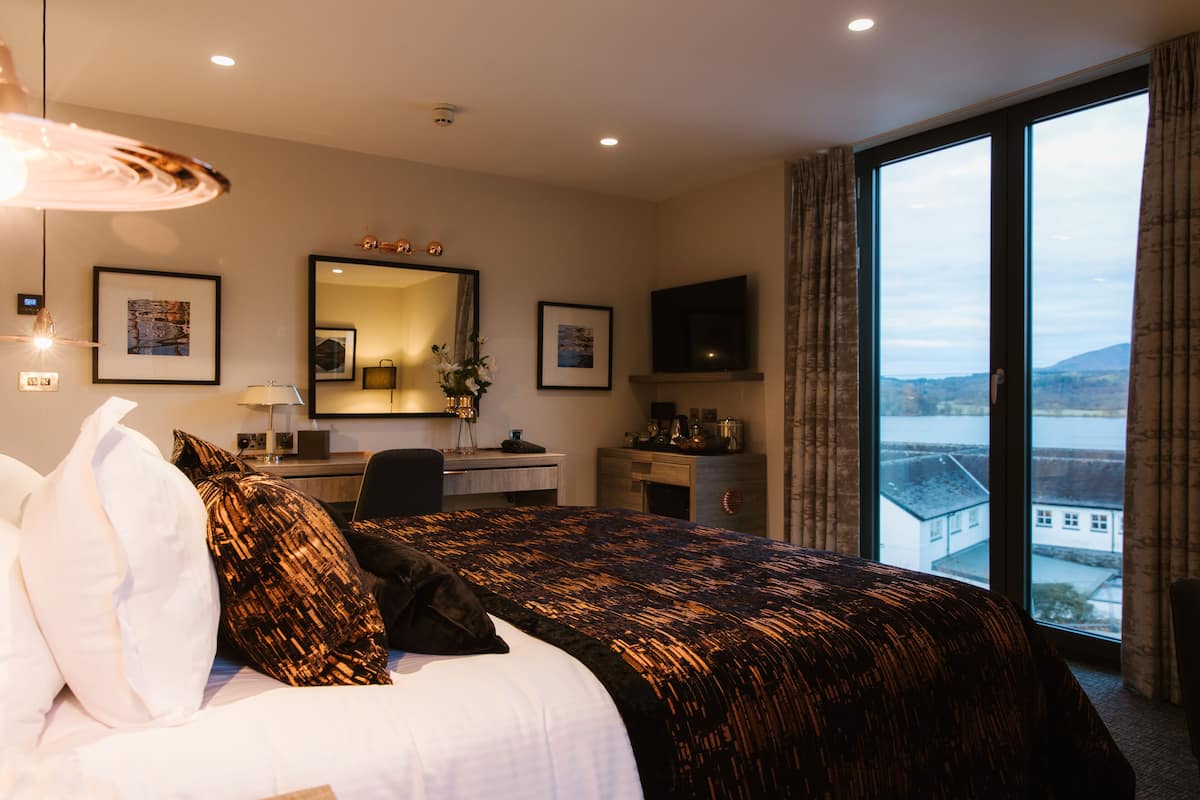 A hotel room with a large bed, a desk, a TV, and a view of a cityscape through large windows.