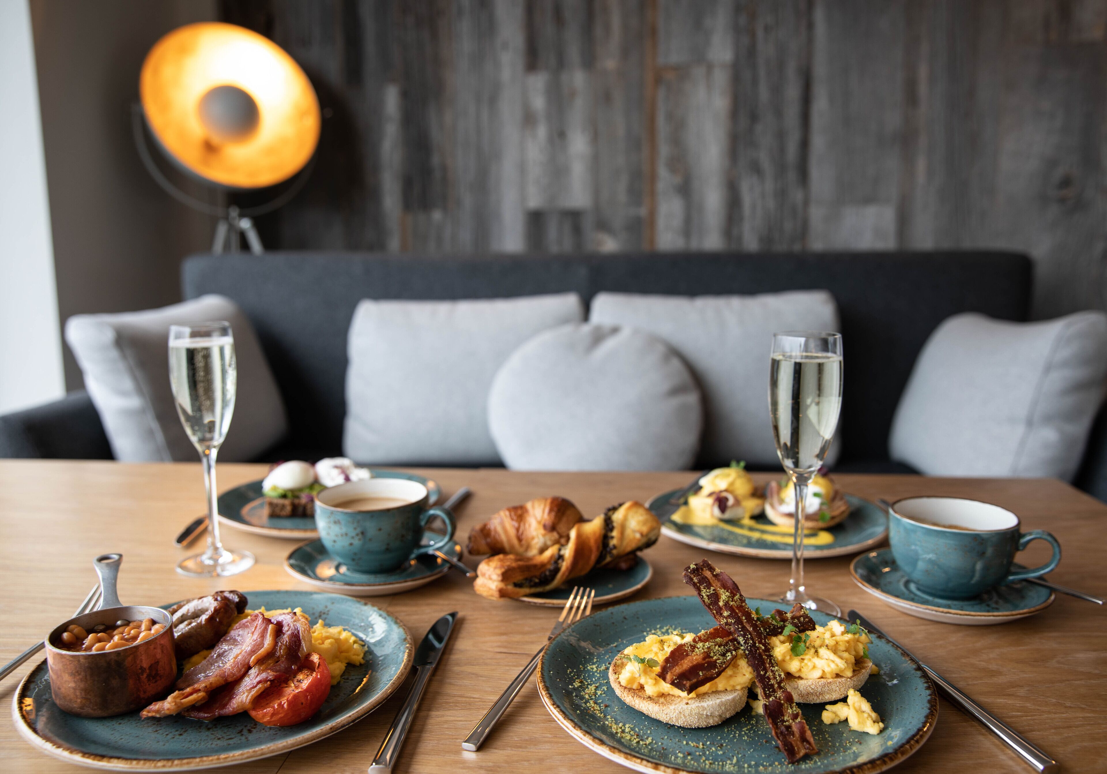A breakfast setting with plates of eggs, bacon, and toast, accompanied by croissants and coffee cups.