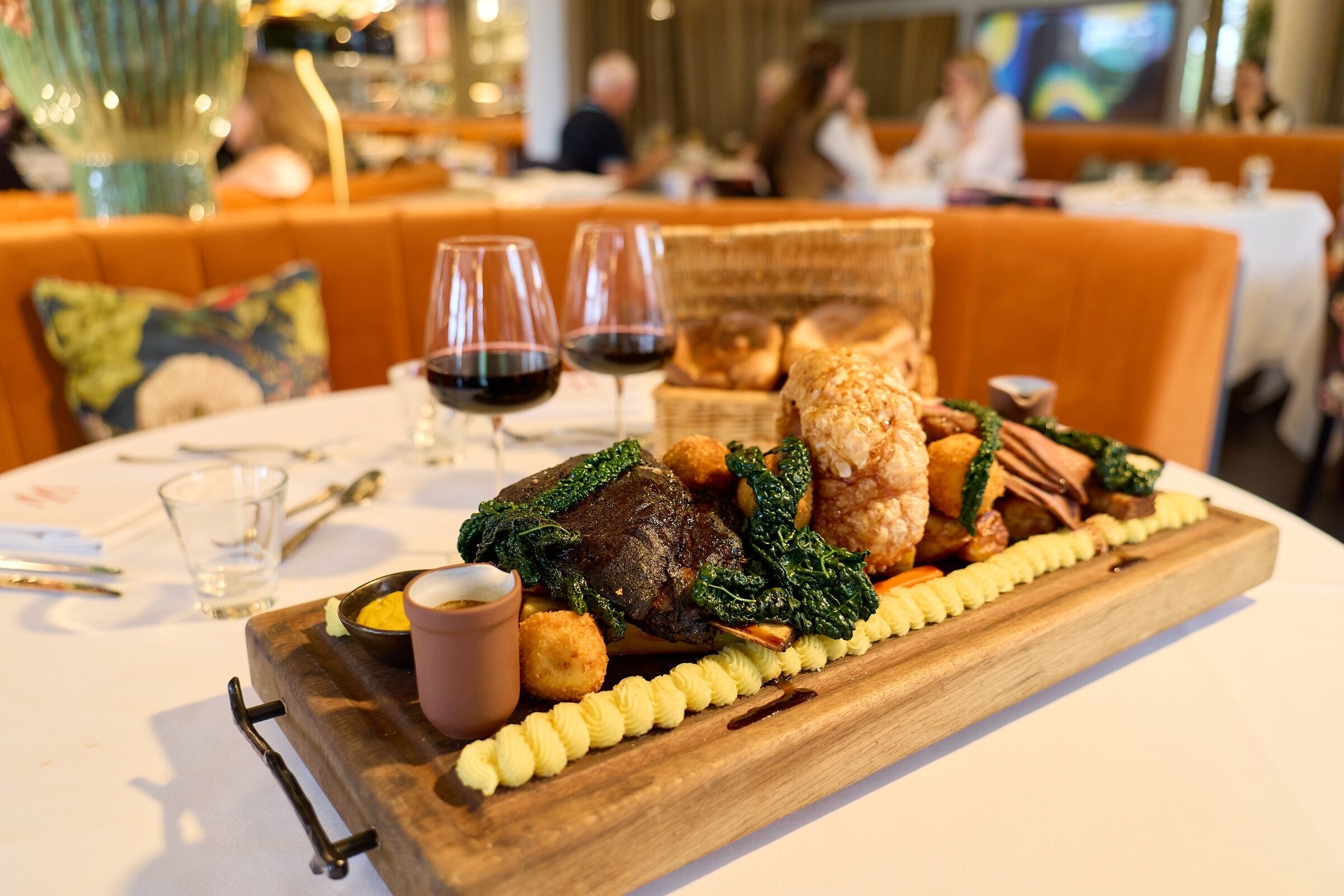 A dining table with a wooden board holding a variety of dishes, including a piece of meat, greens, and other sides. There are two glasses of red wine on the table.