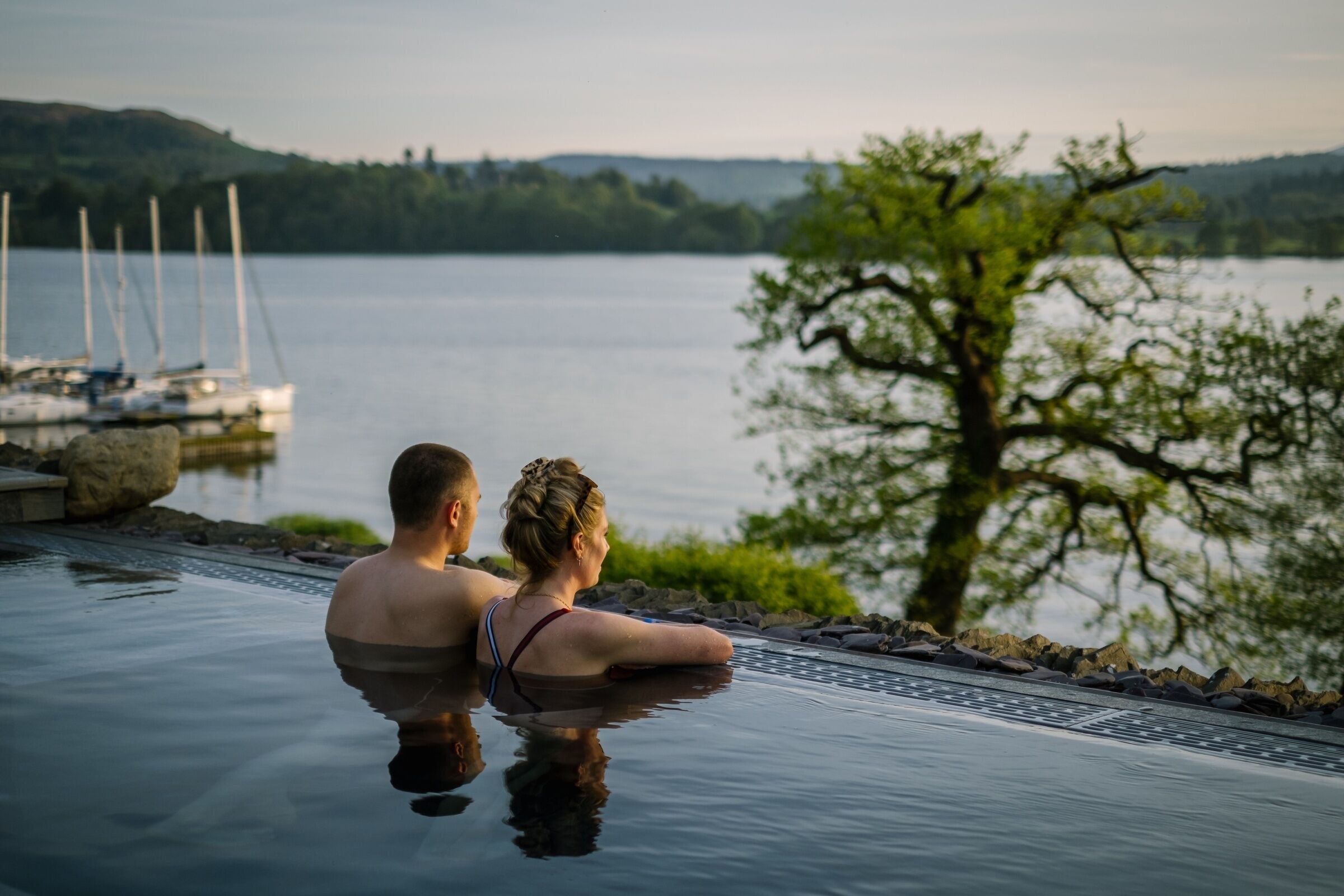 A couple in a private infinity pool overlooking a lake with boats and greenery in the background.