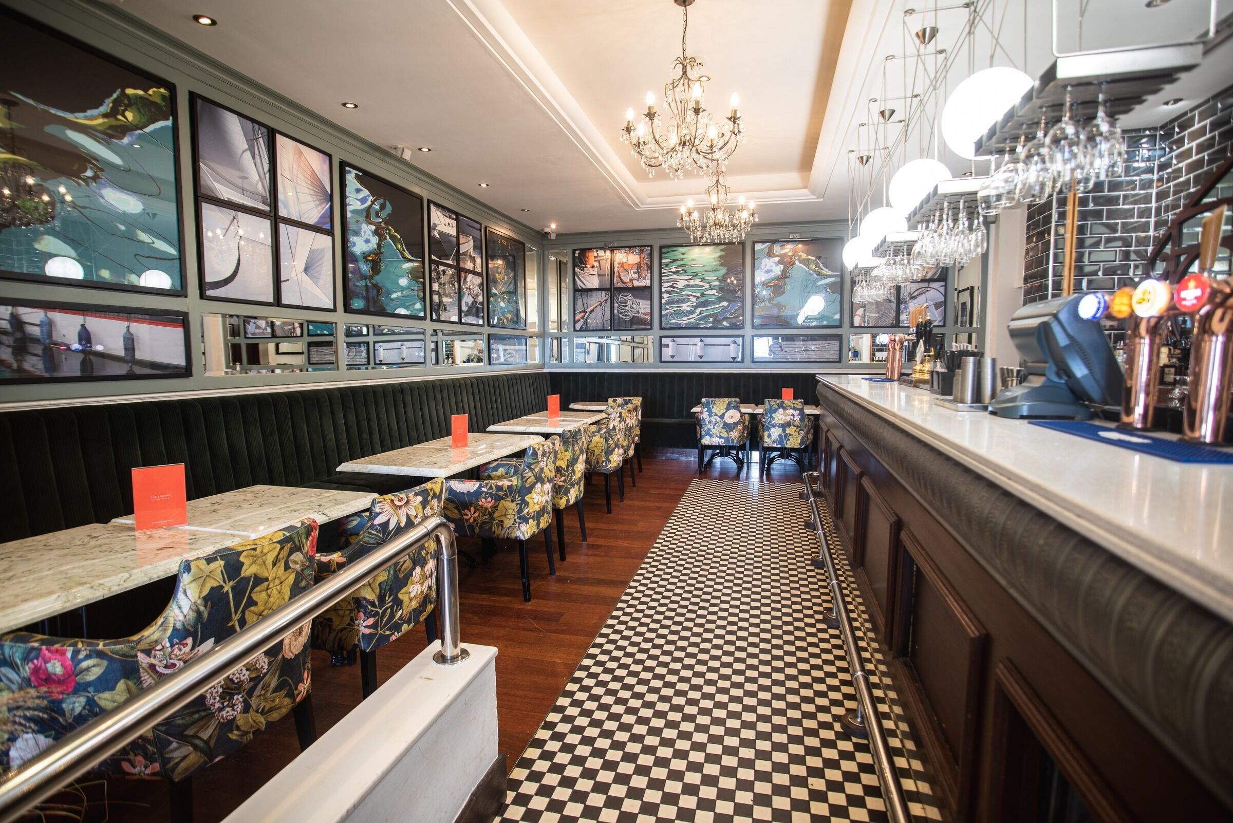 Interior de un restaurante con cabinas, mesas, piso a cuadros y una zona de bar con grifos y vasos.