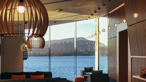 A modern lounge with sofas and ottomans, large windows offering a view of a lake and mountains, and contemporary lighting fixtures. - Low Wood Bay Resort & Spa (Windermere)