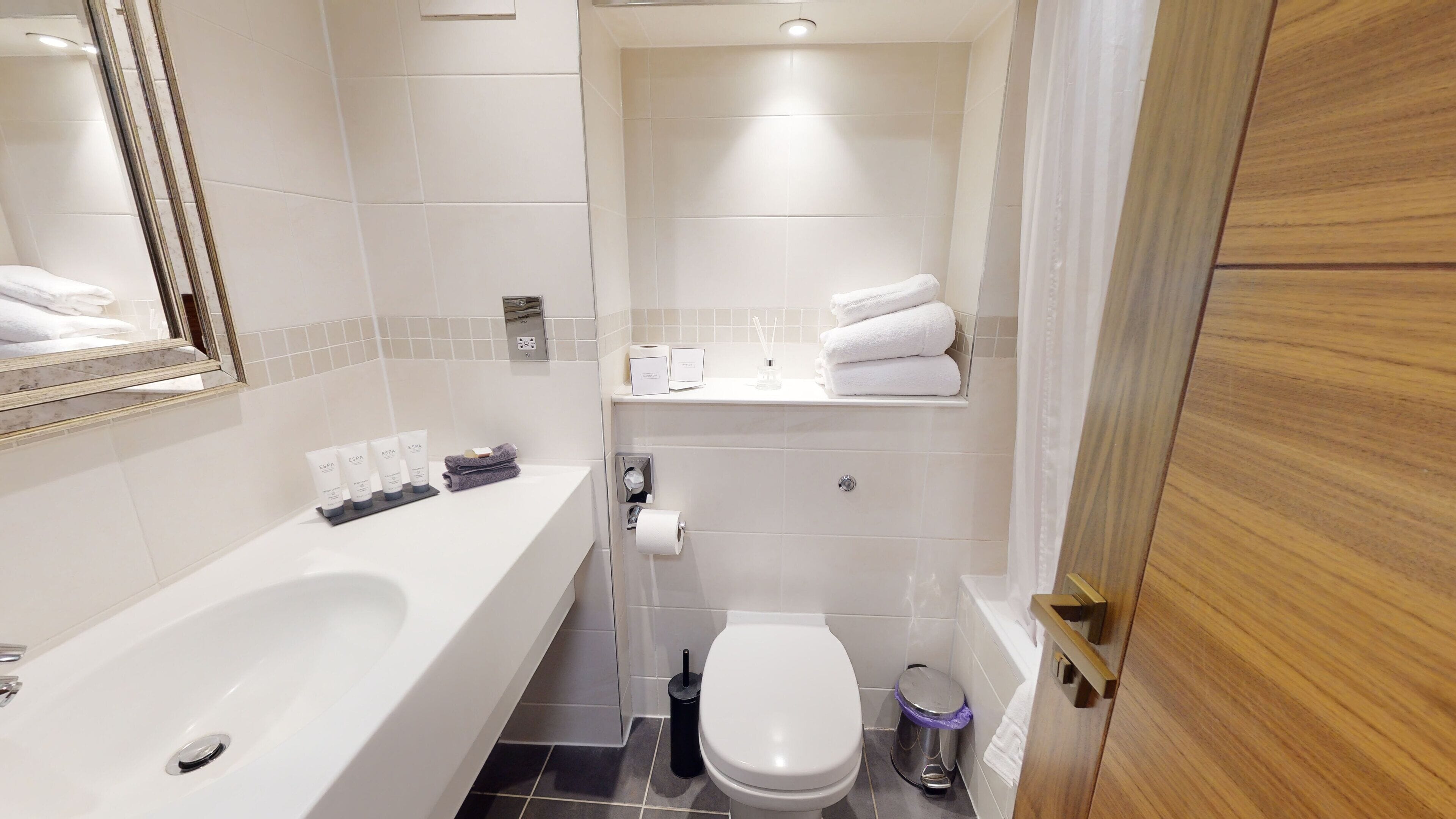 A modern bathroom with a bathtub, toilet, and a shelf with towels and toiletries.