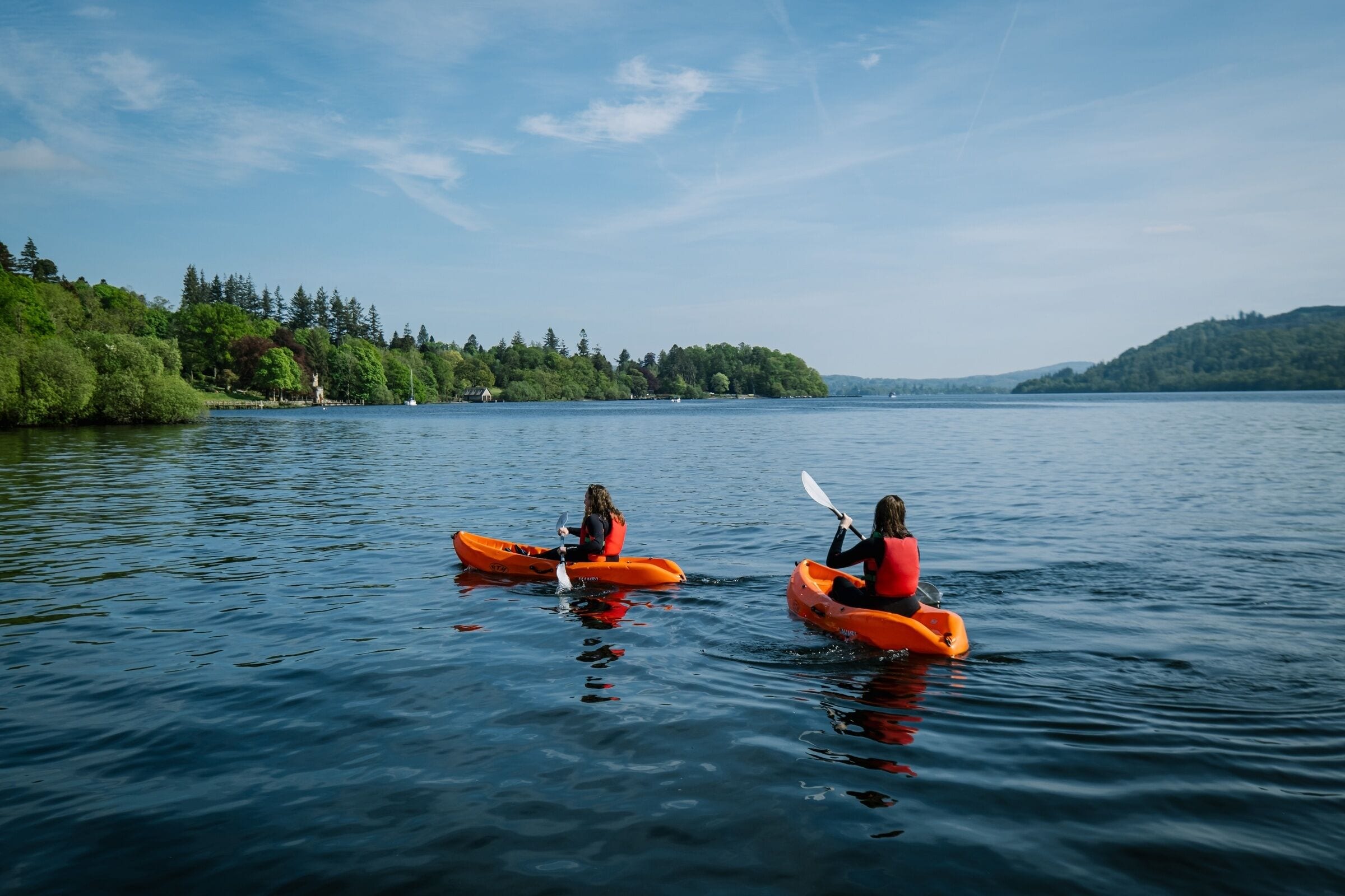 Two kayakers in orange kayaks on a calm lake with a forested shoreline.