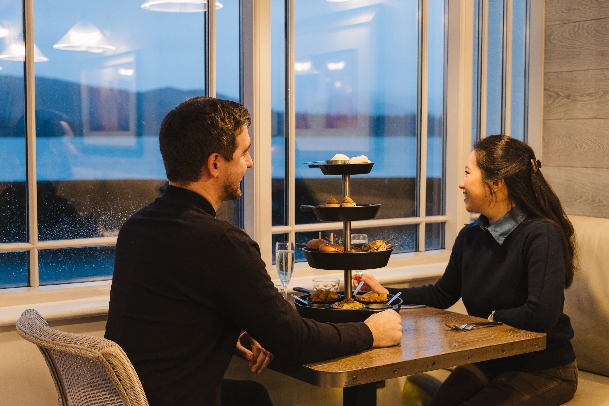 Un coin repas avec une table dressée pour deux personnes, un présentoir à trois étages pour les desserts et une vue sur l’extérieur grâce à de grandes fenêtres.