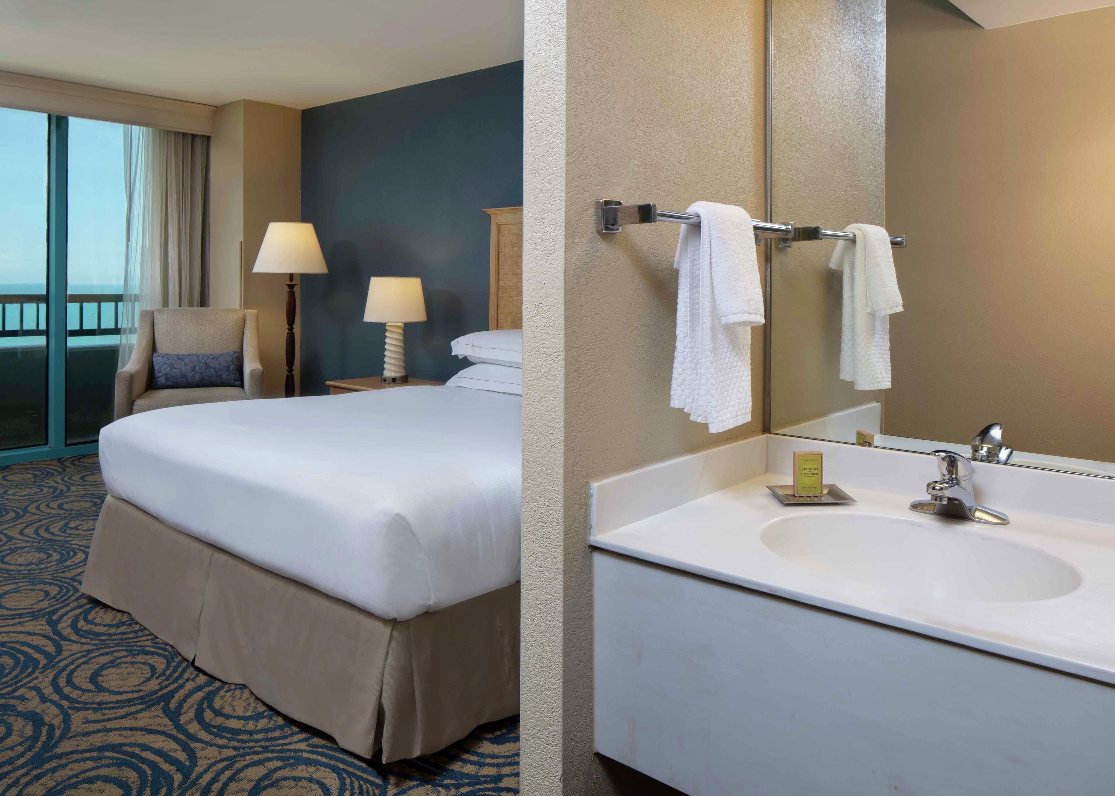 Une chambre d’hôtel avec un lit, une chaise, une table de chevet avec une lampe, et une salle de bain avec un lavabo et un miroir.