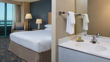 Une chambre d’hôtel avec un lit, une chaise, une table de chevet avec une lampe, et une salle de bain avec un lavabo et un miroir.