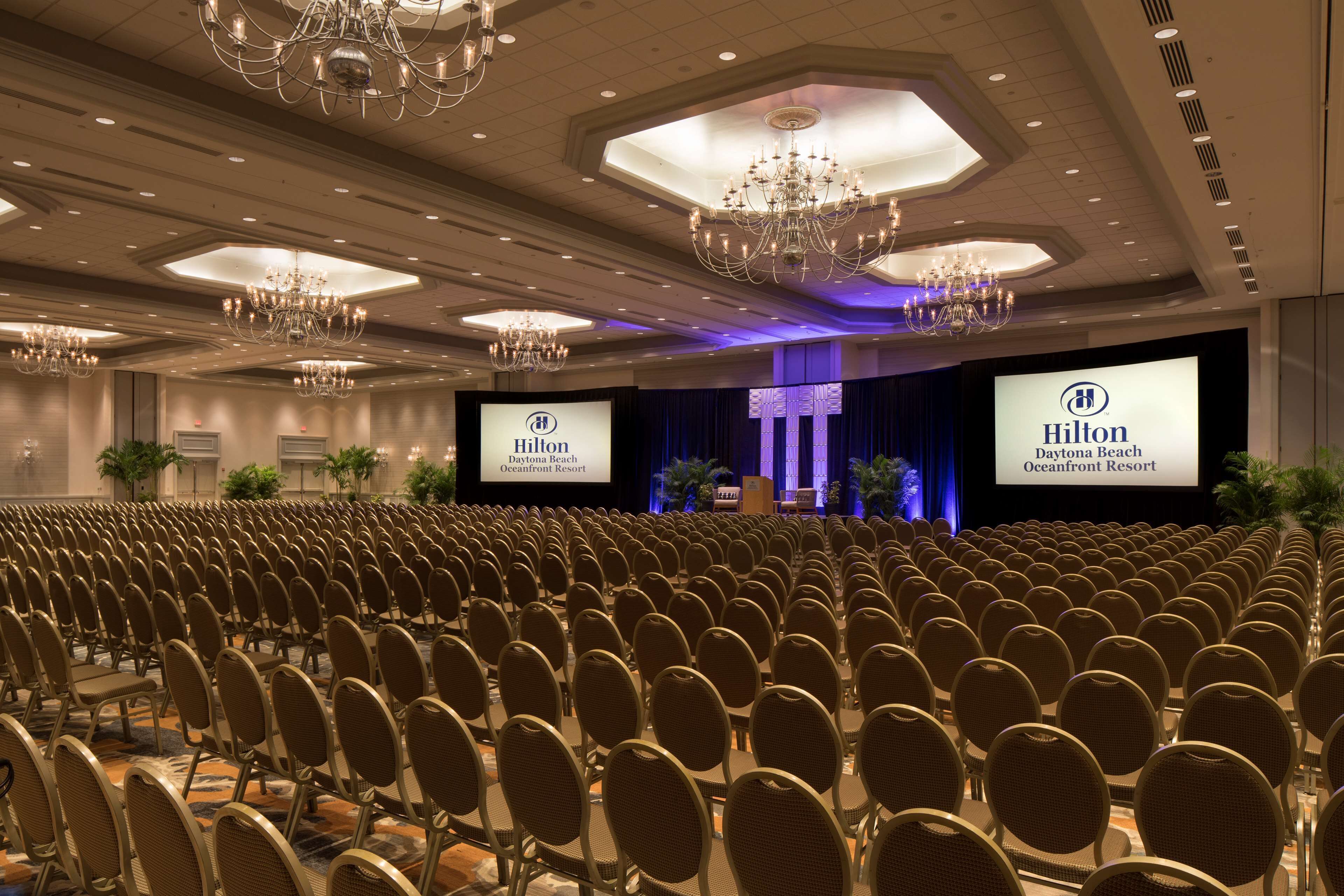 Uma sala de conferências com fileiras de cadeiras, palco com uma tela grande exibindo o logotipo da Hilton e lustres no teto.