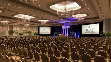 Uma sala de conferências com fileiras de cadeiras, palco com uma tela grande exibindo o logotipo da Hilton e lustres no teto.