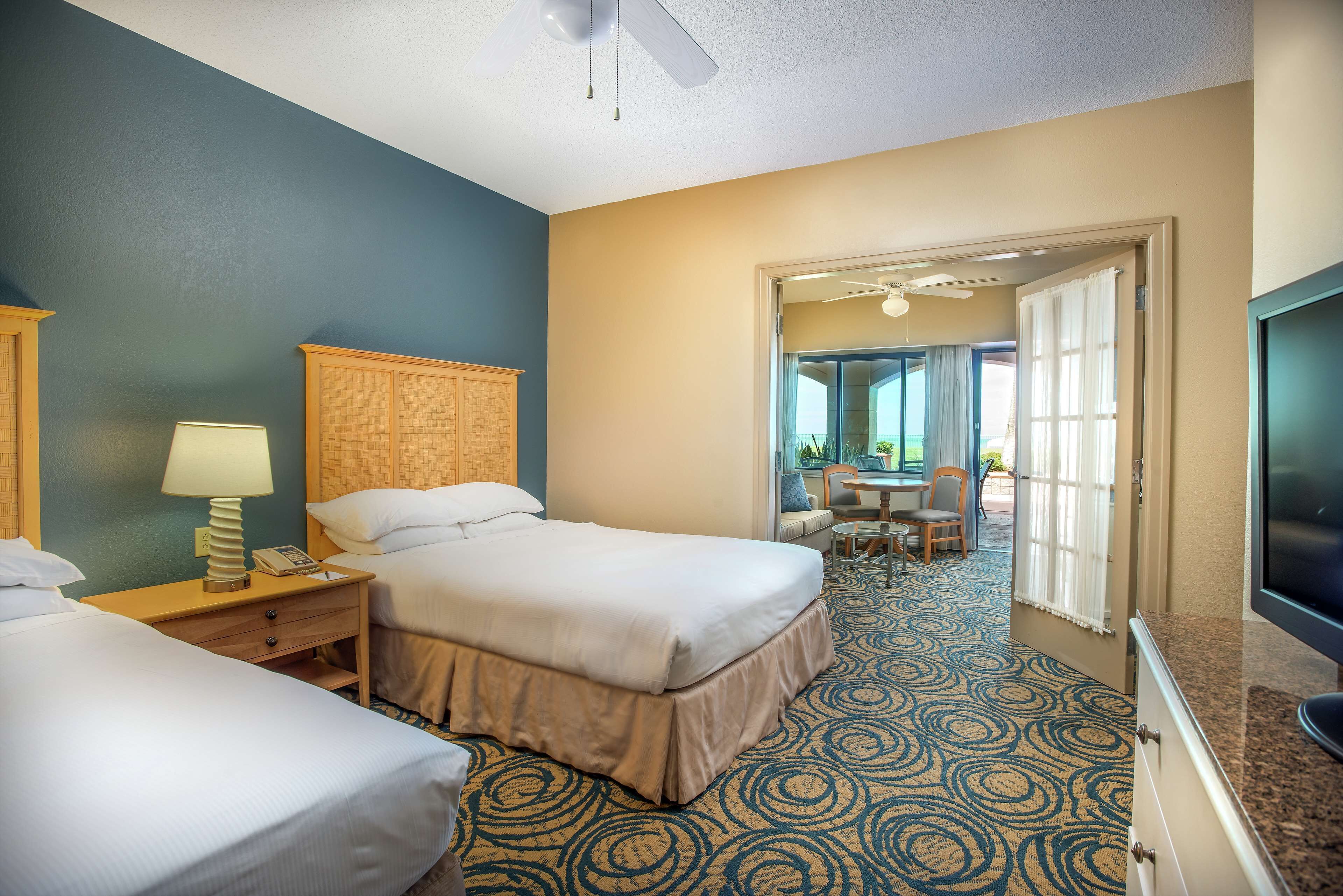 A hotel room with two beds, a desk with a lamp, a television, and a view of the outdoor patio area.