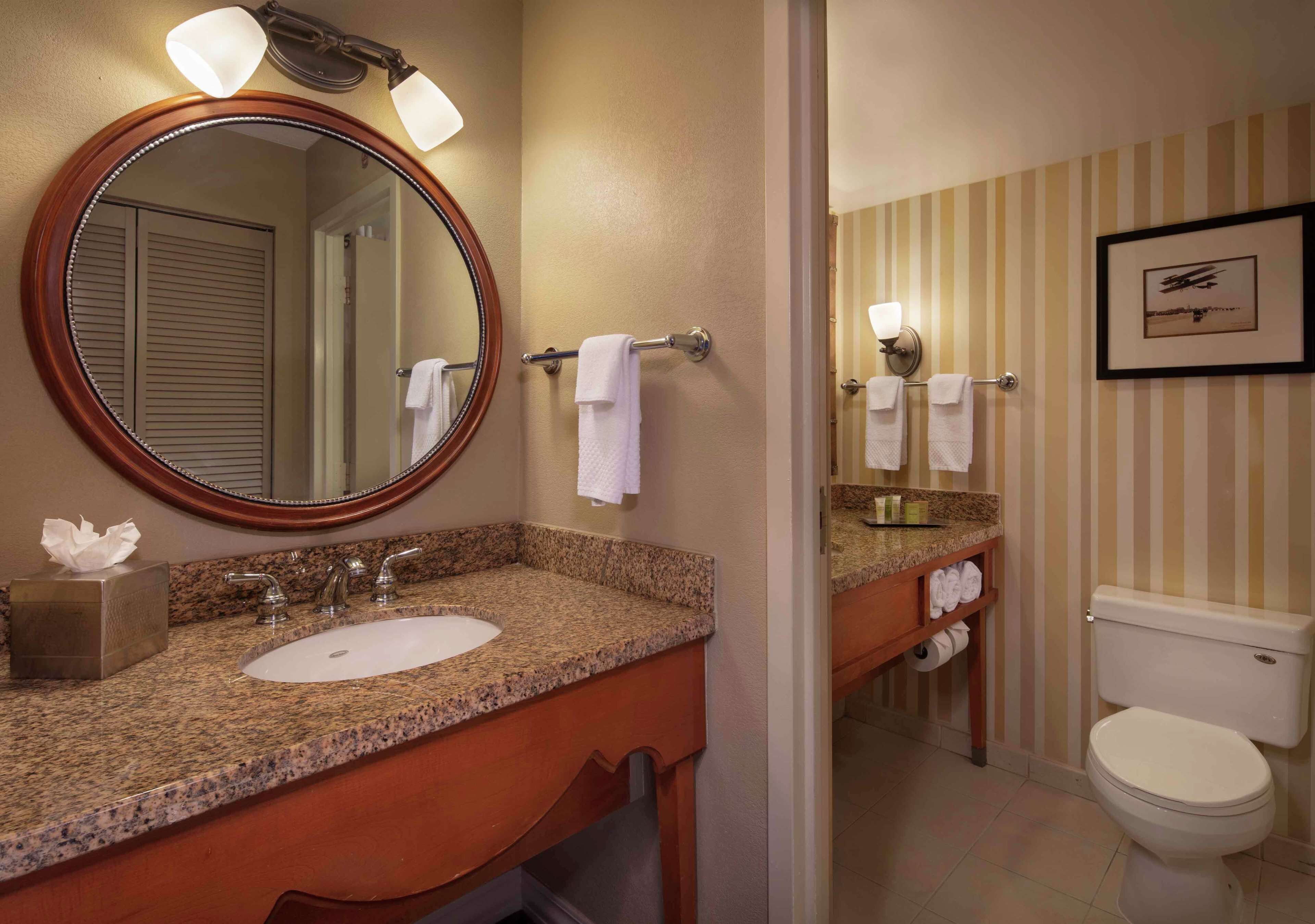 A bathroom with a granite countertop sink, a mirror with lights, a towel rack, a toilet, and a framed picture on the wall.