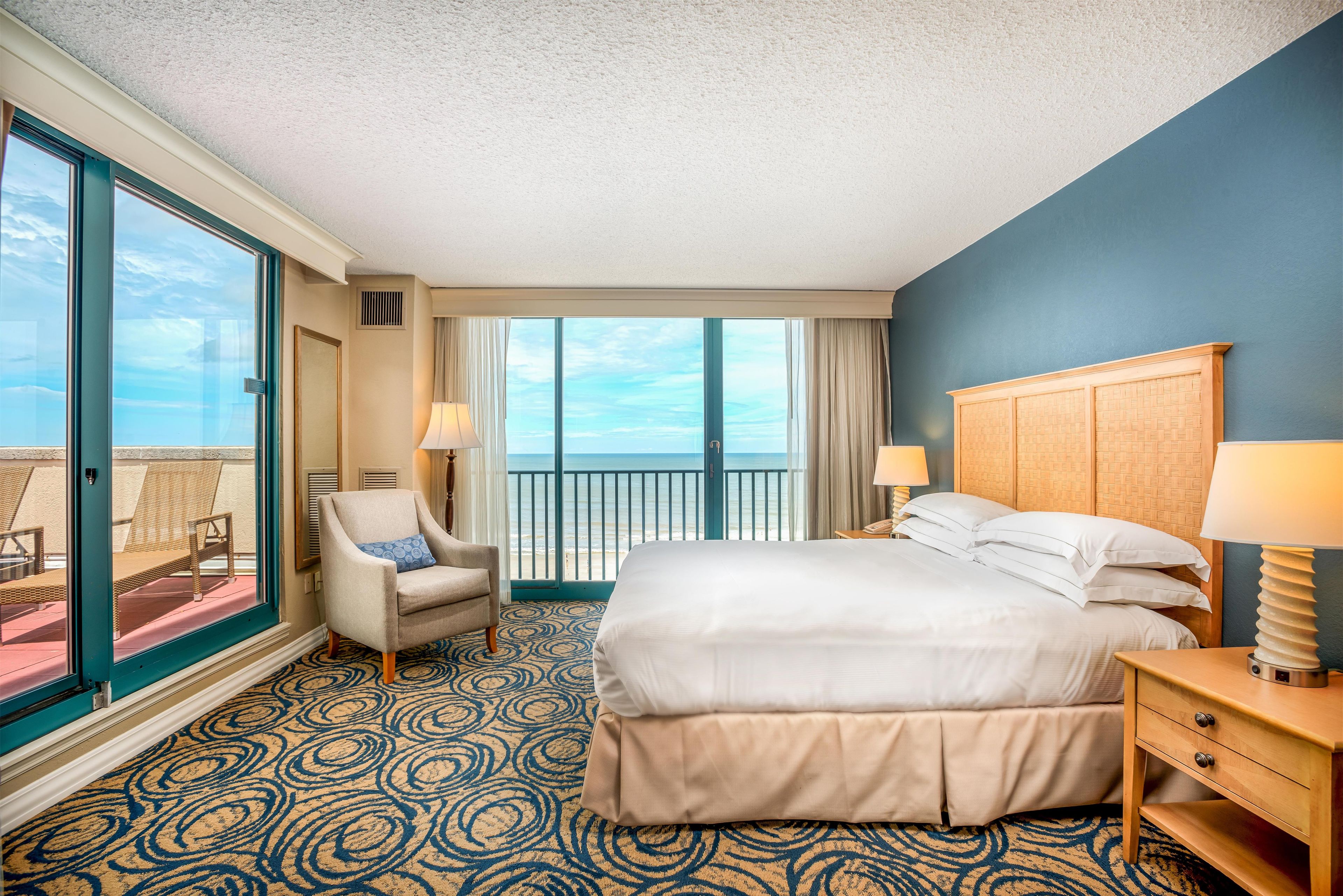 A hotel room with a bed, a chair, a nightstand, a lamp, and a view of the ocean through large windows.