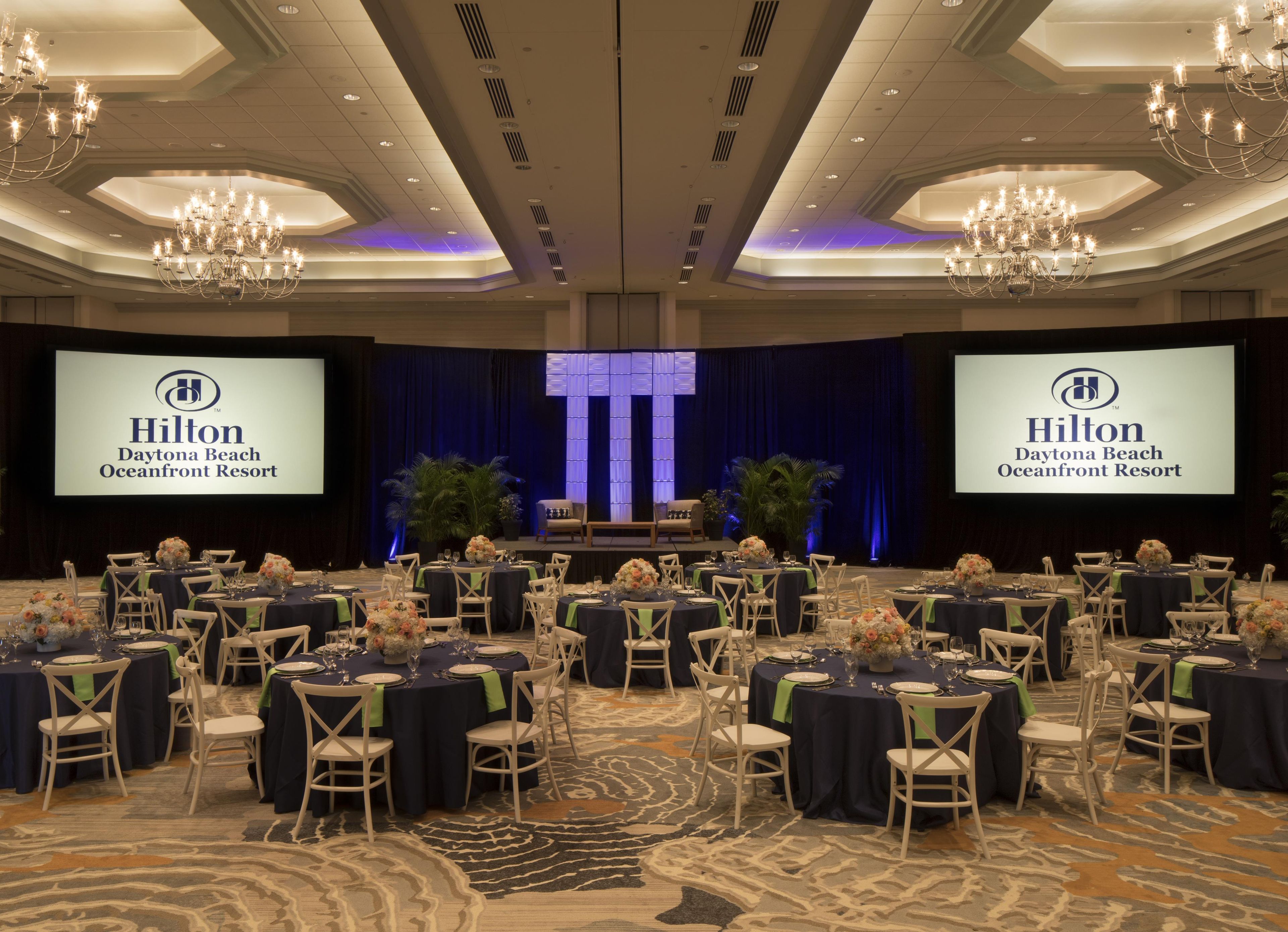 Una sala conferenze all'Hilton Daytona Beach Oceanfront Resort con diversi tavoli apparecchiati per una riunione, un palco con un leggio e un grande schermo che mostra il nome del resort.