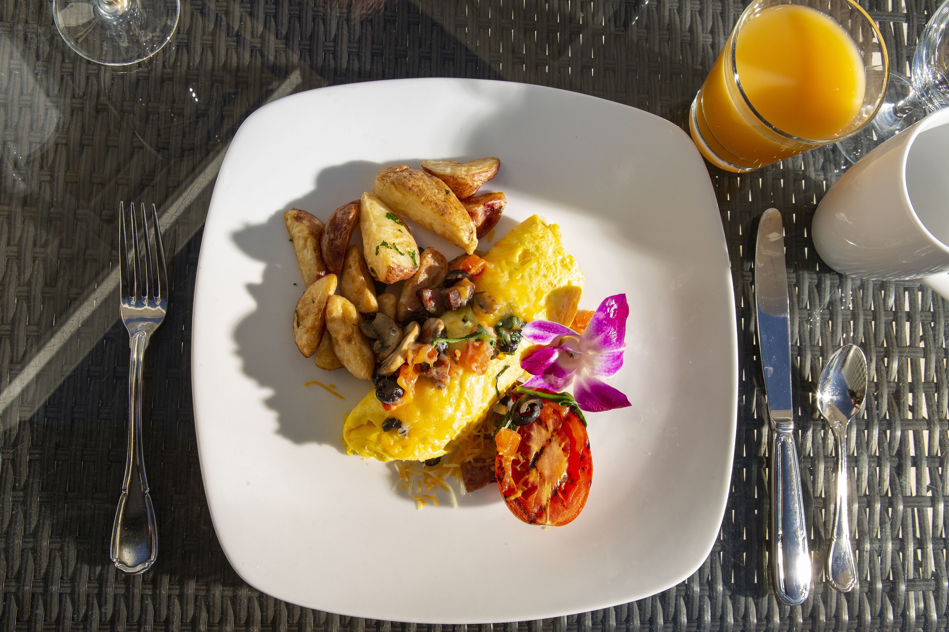 A breakfast plate with eggs, potatoes, and a tomato, accompanied by a glass of orange juice and cutlery on a woven table.