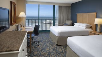 A hotel room with a large bed, a smaller bed, a desk with a lamp, a chair, and a view of the ocean.