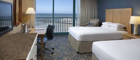 A hotel room with a large bed, a smaller bed, a desk with a lamp, a chair, and a view of the ocean.