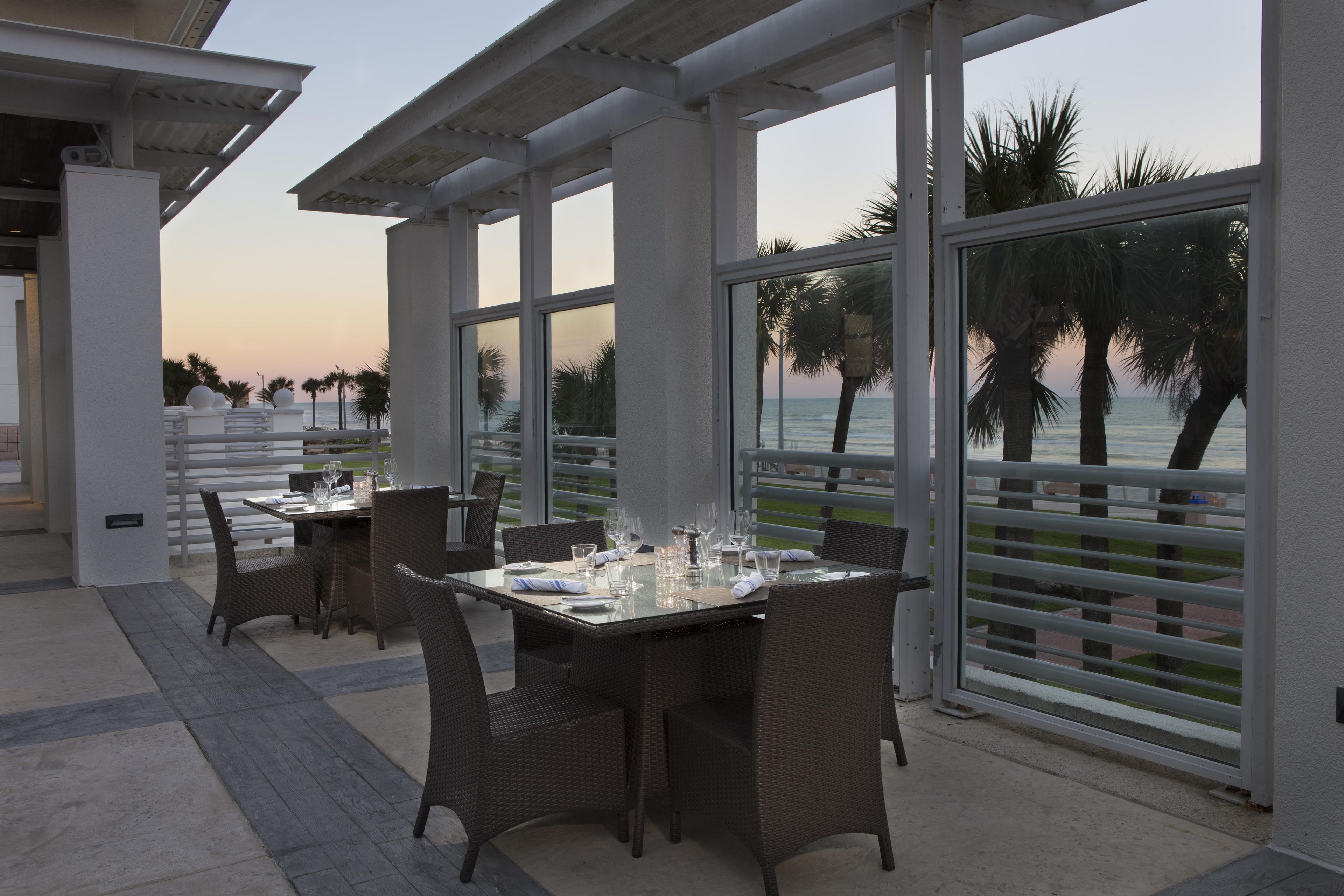 Un restaurante con terraza al aire libre que da al mar, con mesas y sillas y vistas a palmeras.