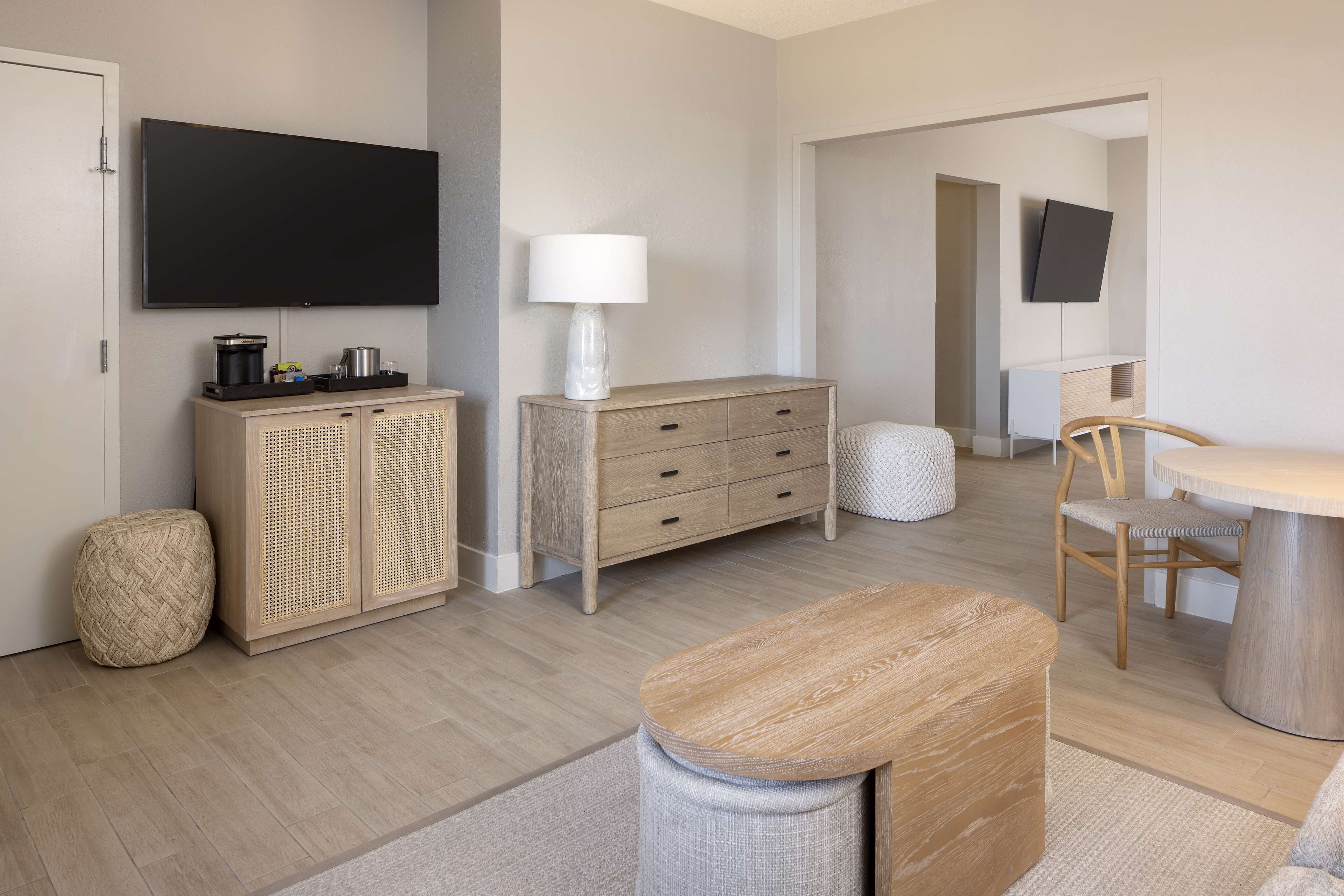 A modern living room with a flat-screen TV mounted on the wall, a wooden cabinet with drawers, a round wooden table, and a comfortable chair.