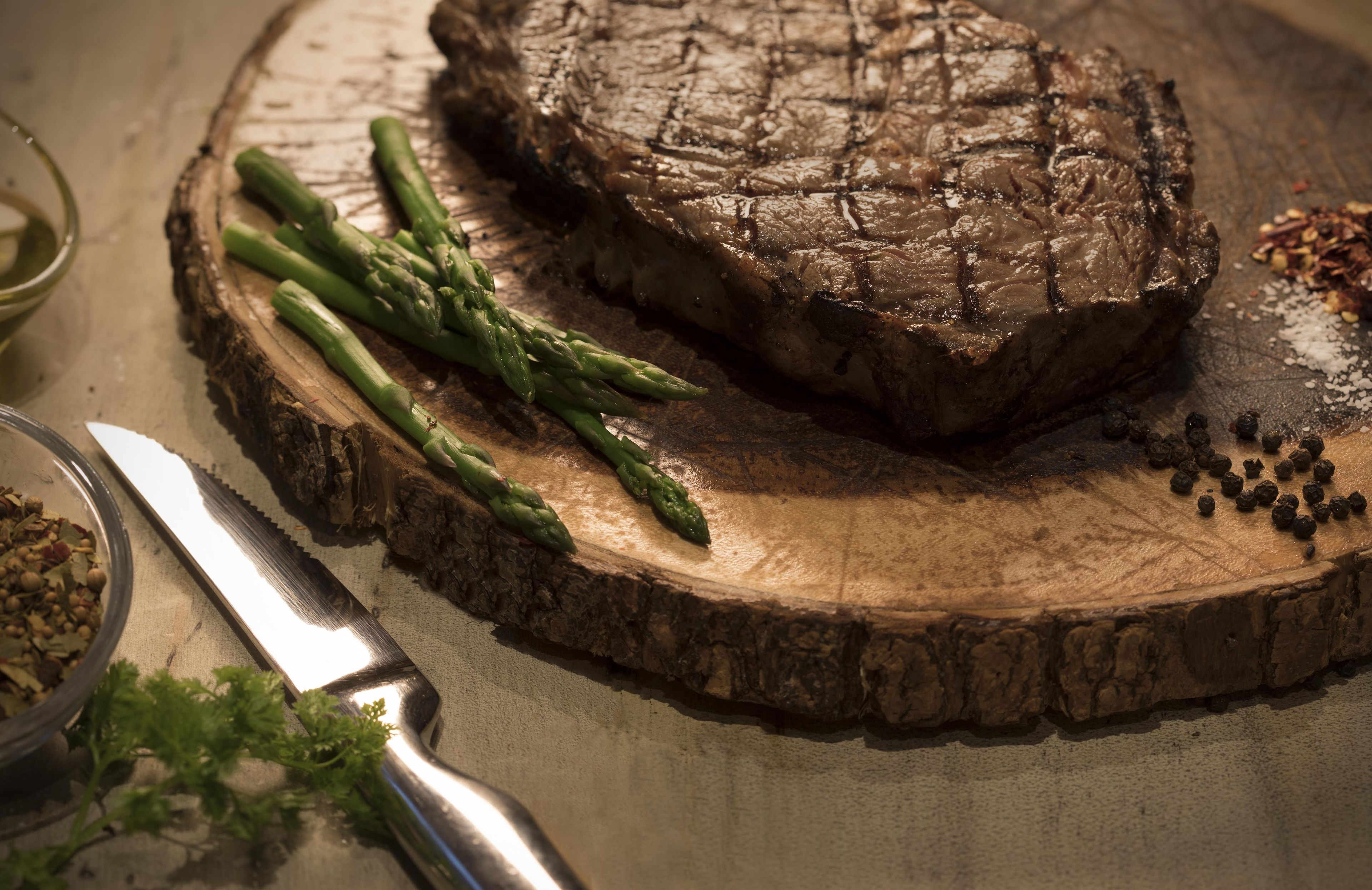 Un steak servi sur une planche en bois, accompagné d’asperges et de grains de poivre.