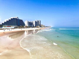 A beach with clear waters, a sandy shore, and a row of hotels in the background.