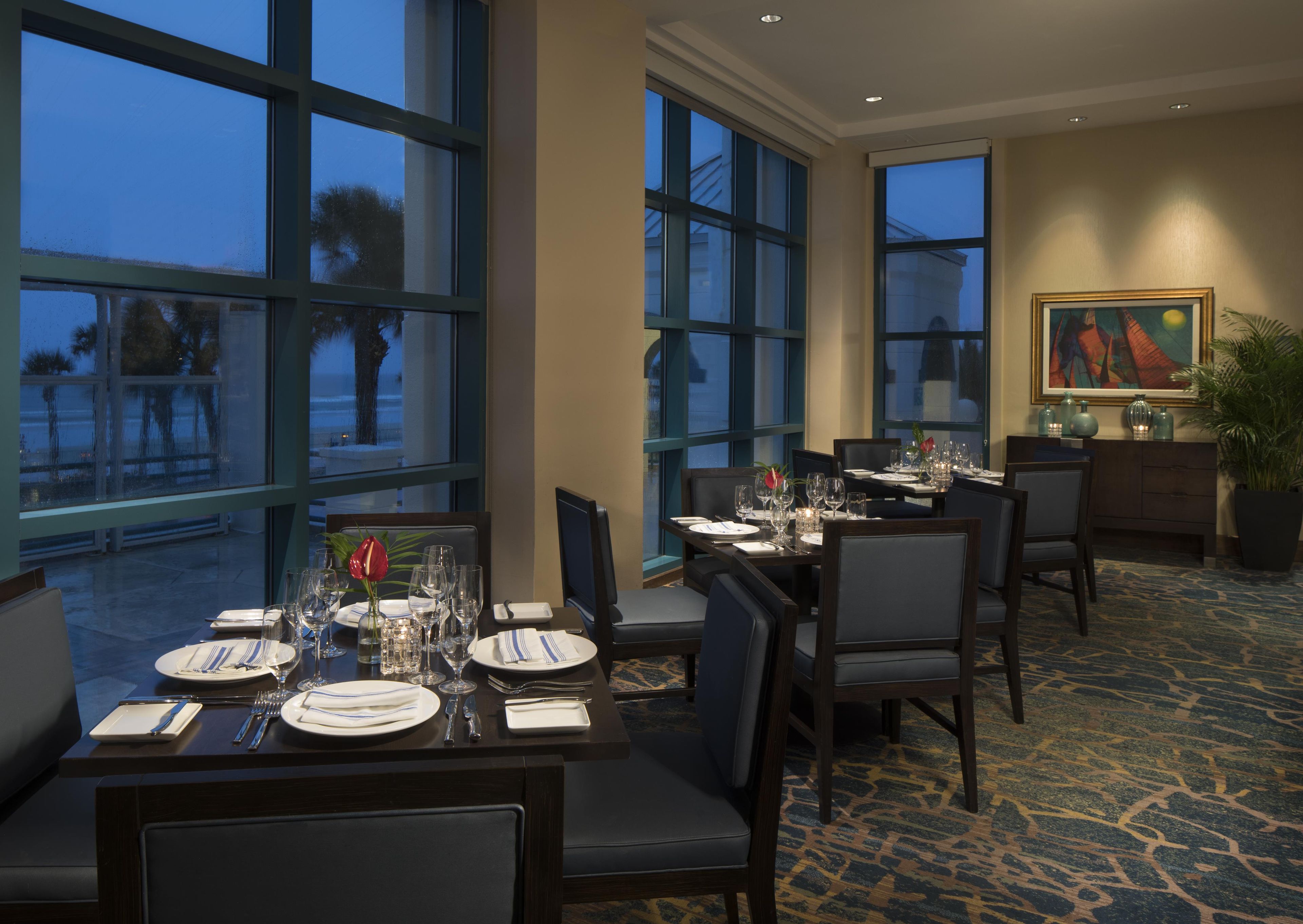 A dining area with tables set for four, large windows, and a painting on the wall.