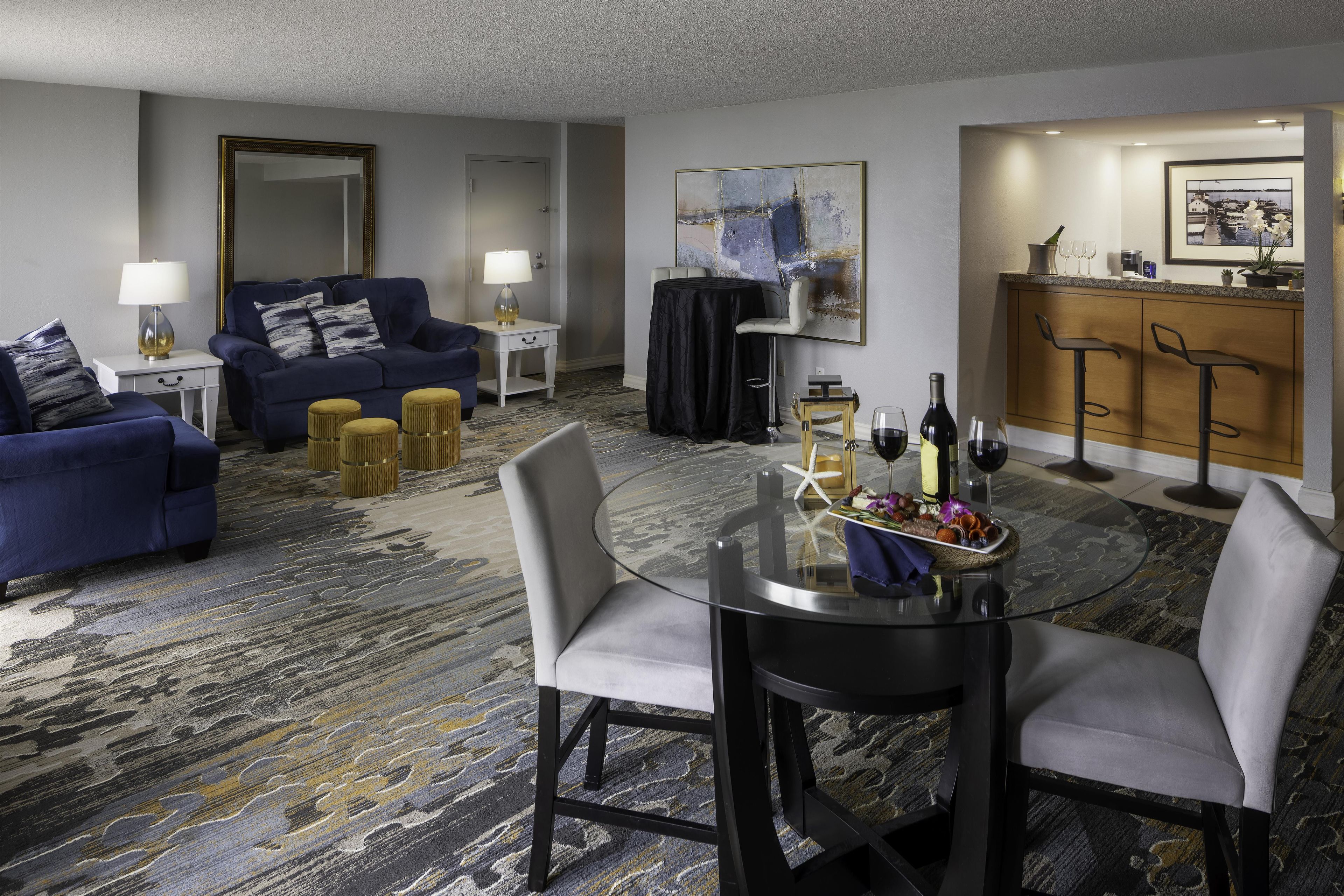 A hotel room with a seating area, a dining table with chairs, a bar area, and a large window.