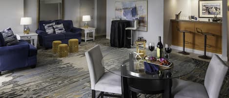 A hotel room with a seating area, a dining table with chairs, a bar area, and a large window.